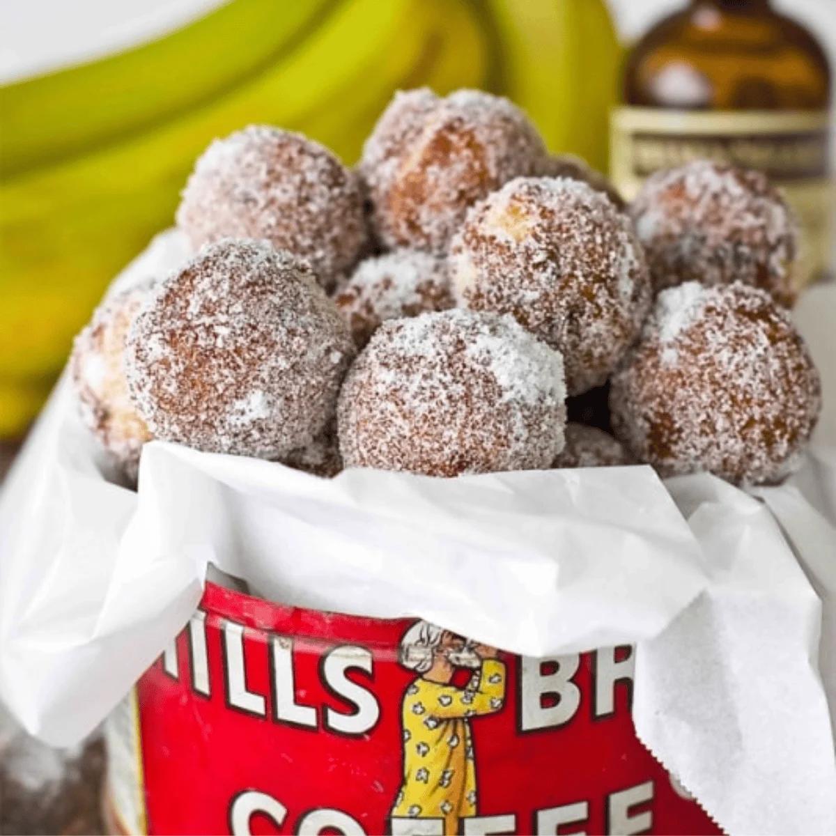 A container filled with powdered sugar-coated doughnut holes, surrounded by bananas and a bottle in the background, brings to mind the delicious treat of 10 Minute Baked Banana Bread Doughnut Holes.
