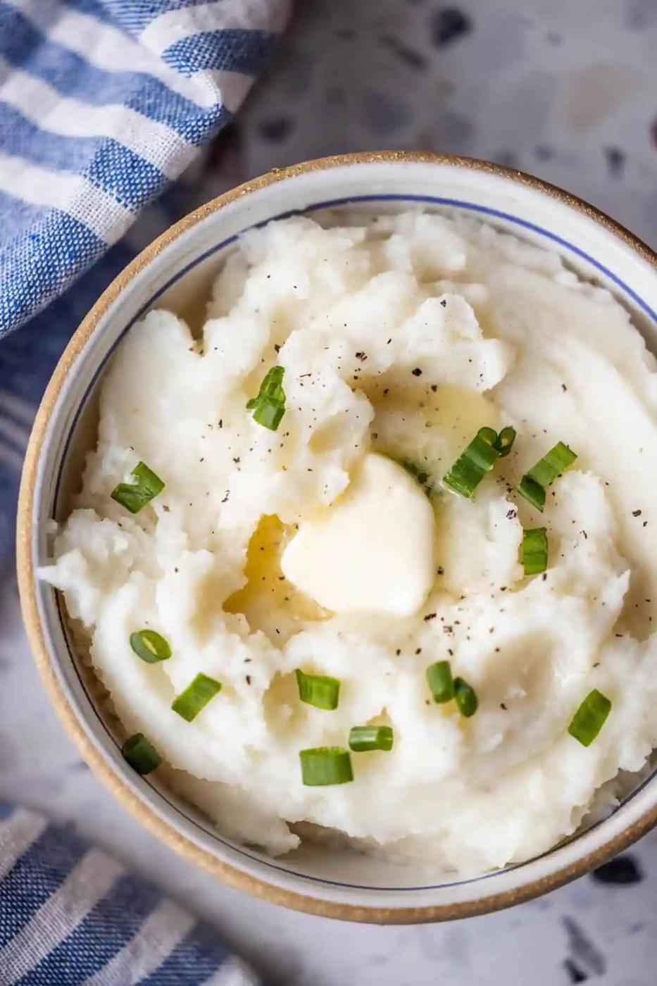A bowl of Instant Pot Mashed Potatoes topped with a pat of butter, chopped green onions, and black pepper sits beside a striped cloth.