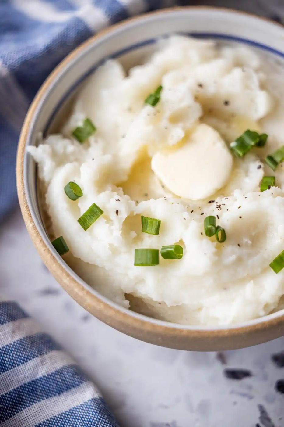 Bowl of Instant Pot Mashed Potatoes topped with a pat of butter, chopped green onions, and black pepper, with a blue and white striped cloth nearby.