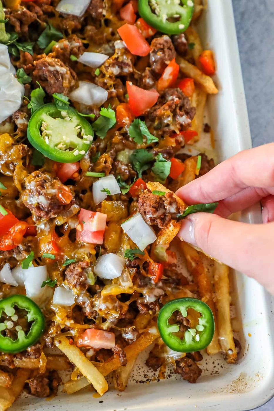 A hand picks up loaded nacho fries, topped with ground beef, melted cheese, diced tomatoes, onions, jalapeños, and cilantro from a tray—perfect for those craving easy nacho fries packed with bold flavor.
