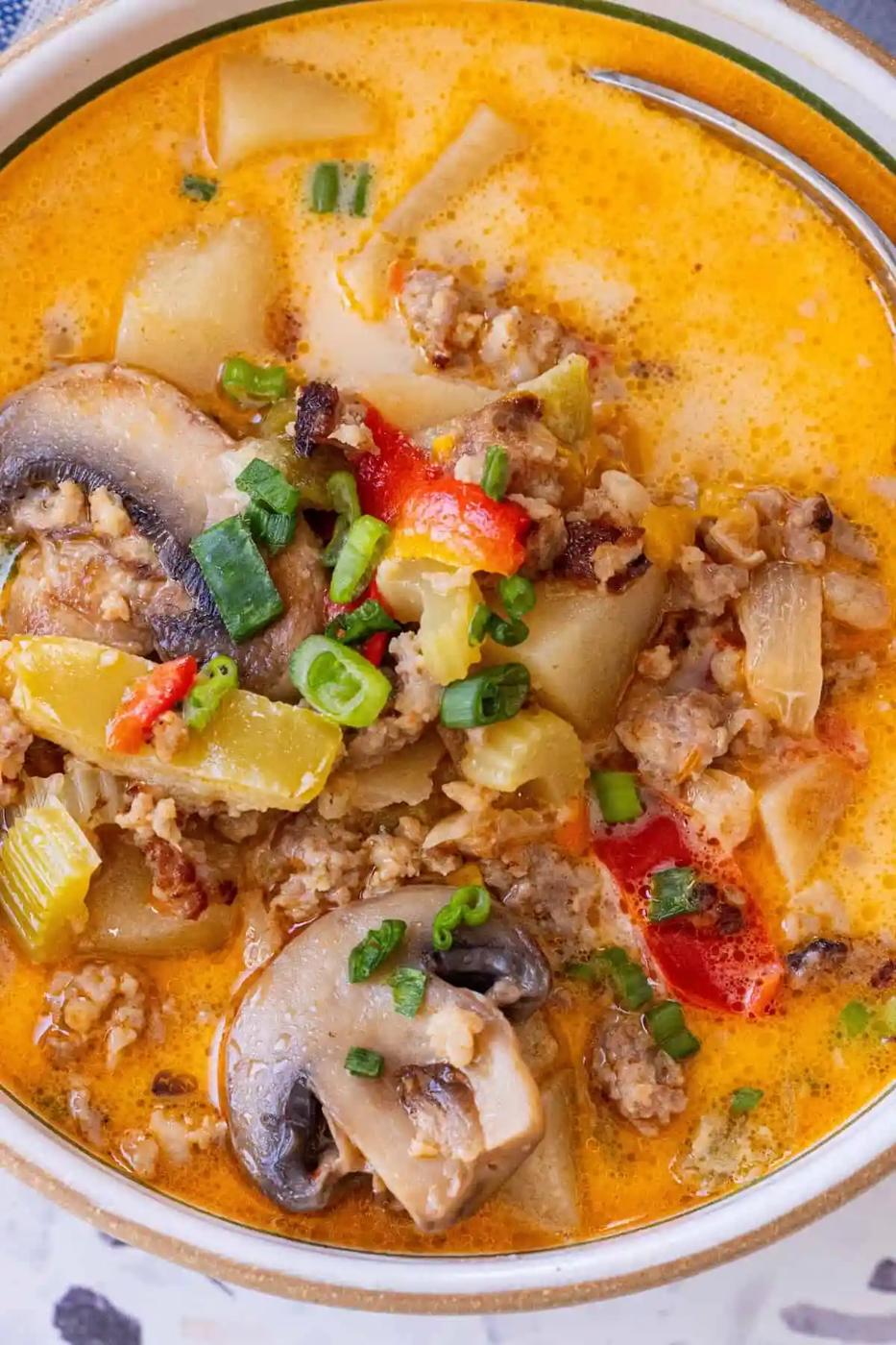 A bowl of creamy orange soup with ground meat, sliced mushrooms, potatoes, bell peppers, and topped with chopped green onions—just like our 20 Minute Creamy Sausage and Mushroom Soup for a quick and hearty meal.