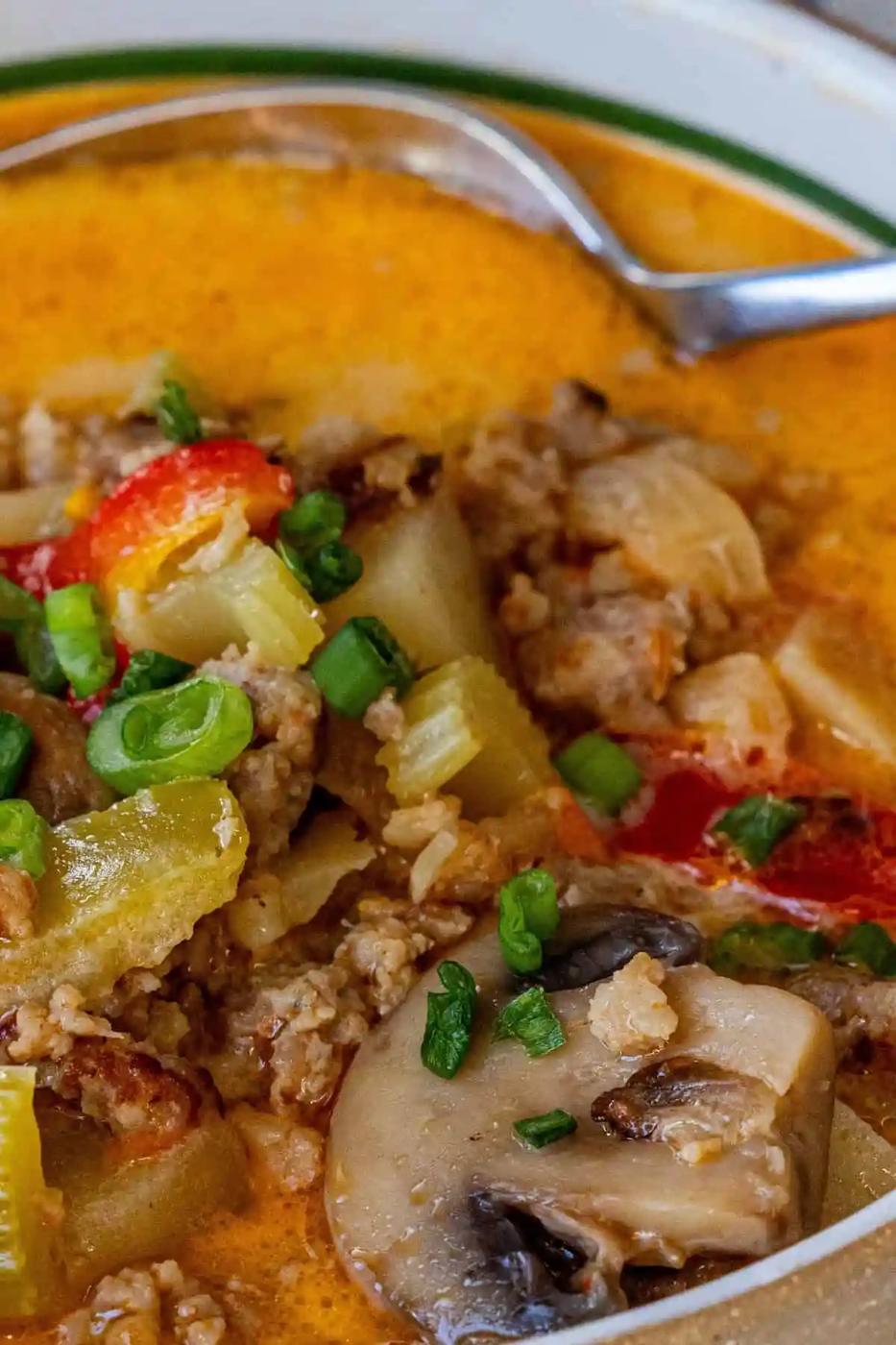 Close-up of a bowl of low carb mushroom sausage chowder with ground meat, mushrooms, green onions, red peppers, celery, and potatoes in a creamy orange broth. A spoon rests in the bowl.