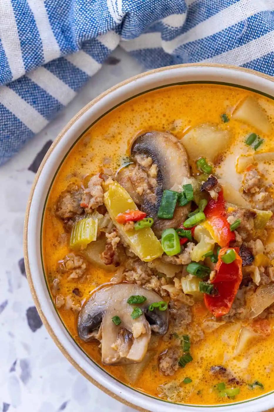 A bowl of creamy one pot mushroom soup sausage with ground meat, red bell peppers, celery, potatoes, and green onions, placed on a light surface next to a blue and white striped cloth.