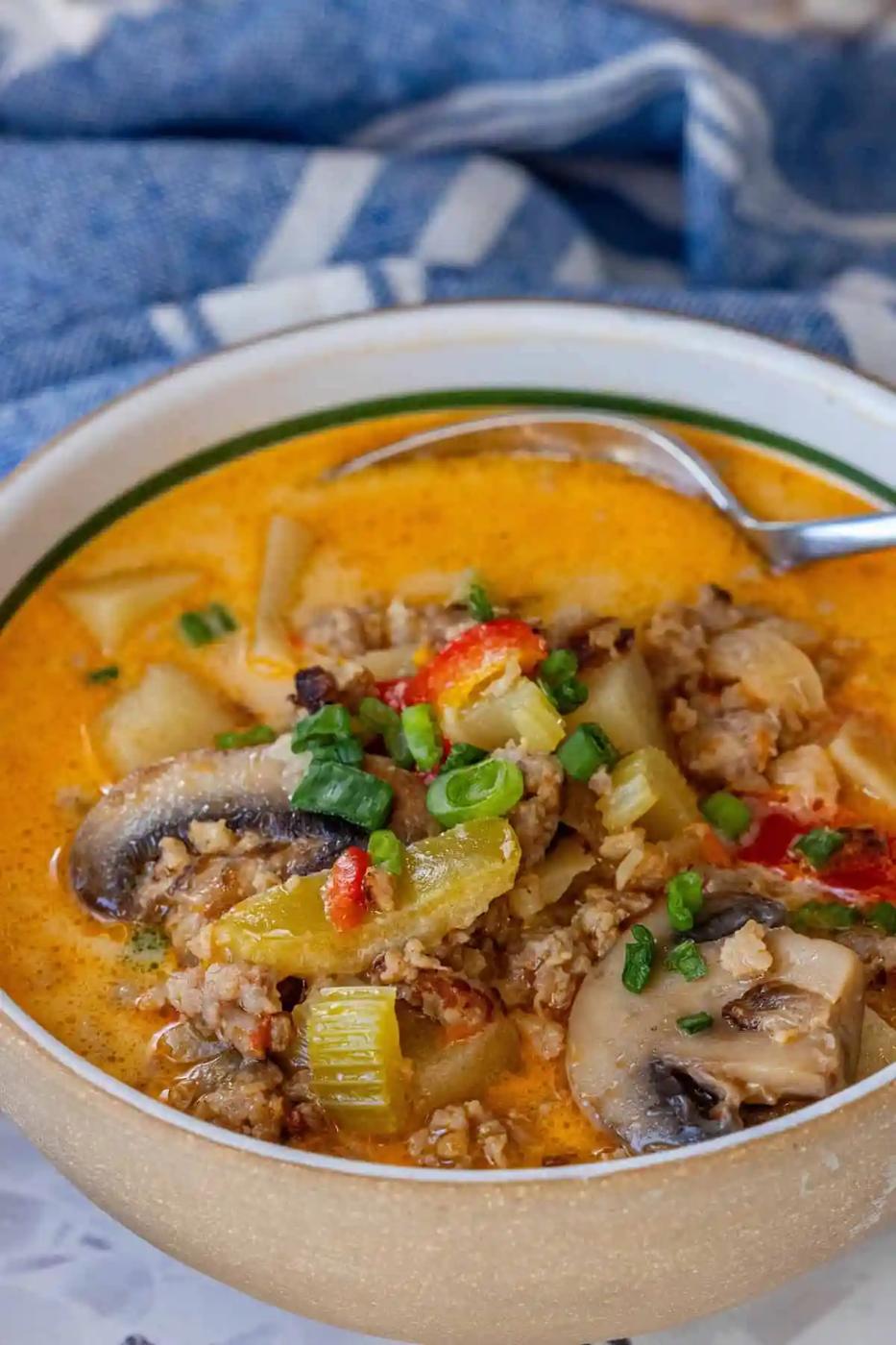 A bowl of Easy Creamy Mushroom and Sausage Soup Recipe featuring ground meat, mushrooms, celery, potatoes, green onions, and red peppers. A spoon rests inside and a blue and white cloth is in the background.