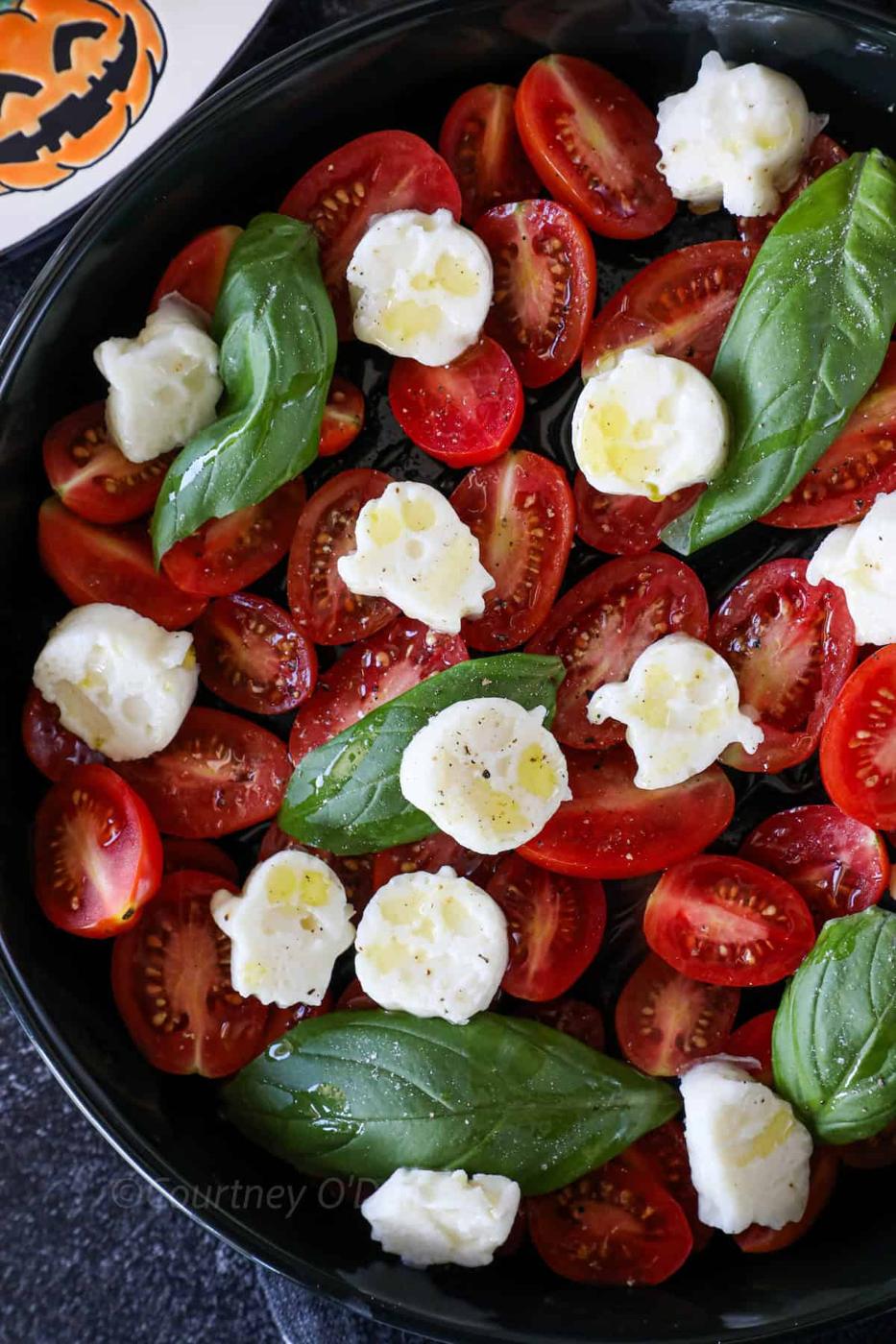 A bowl contains halved cherry tomatoes, fresh basil leaves, and dollops of mozzarella cheese, drizzled with olive oil. A Halloween-themed plate with a pumpkin face design is partly visible.