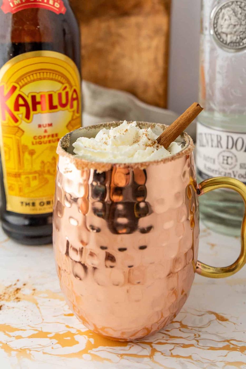 A copper mug filled with a whipped cream-topped beverage, garnished with a cinnamon stick, sits in front of a Kahlúa bottle and a bottle of Silver Dollar vodka.