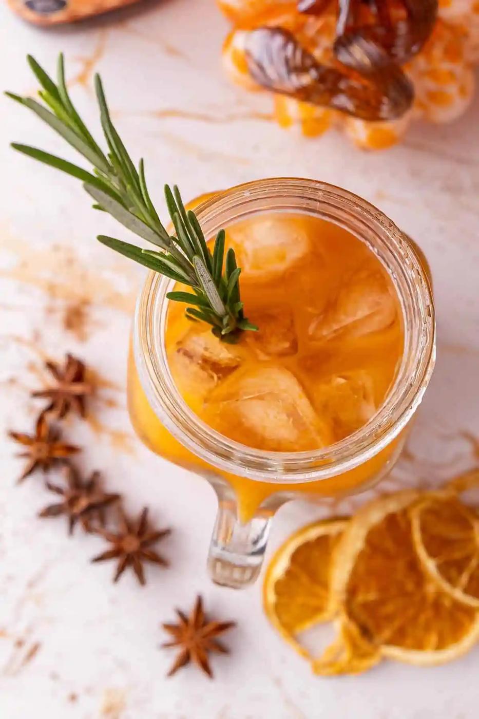 A glass mug with iced orange tea garnished with rosemary, surrounded by star anise and dried orange slices.