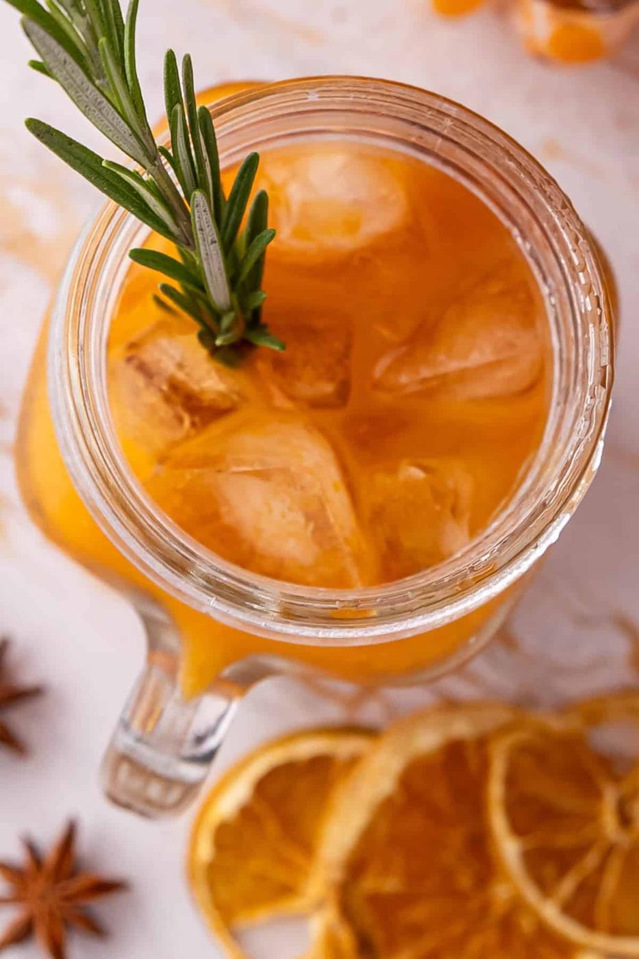A glass mug filled with orange iced tea, garnished with rosemary, surrounded by star anise and dried orange slices on a light surface.