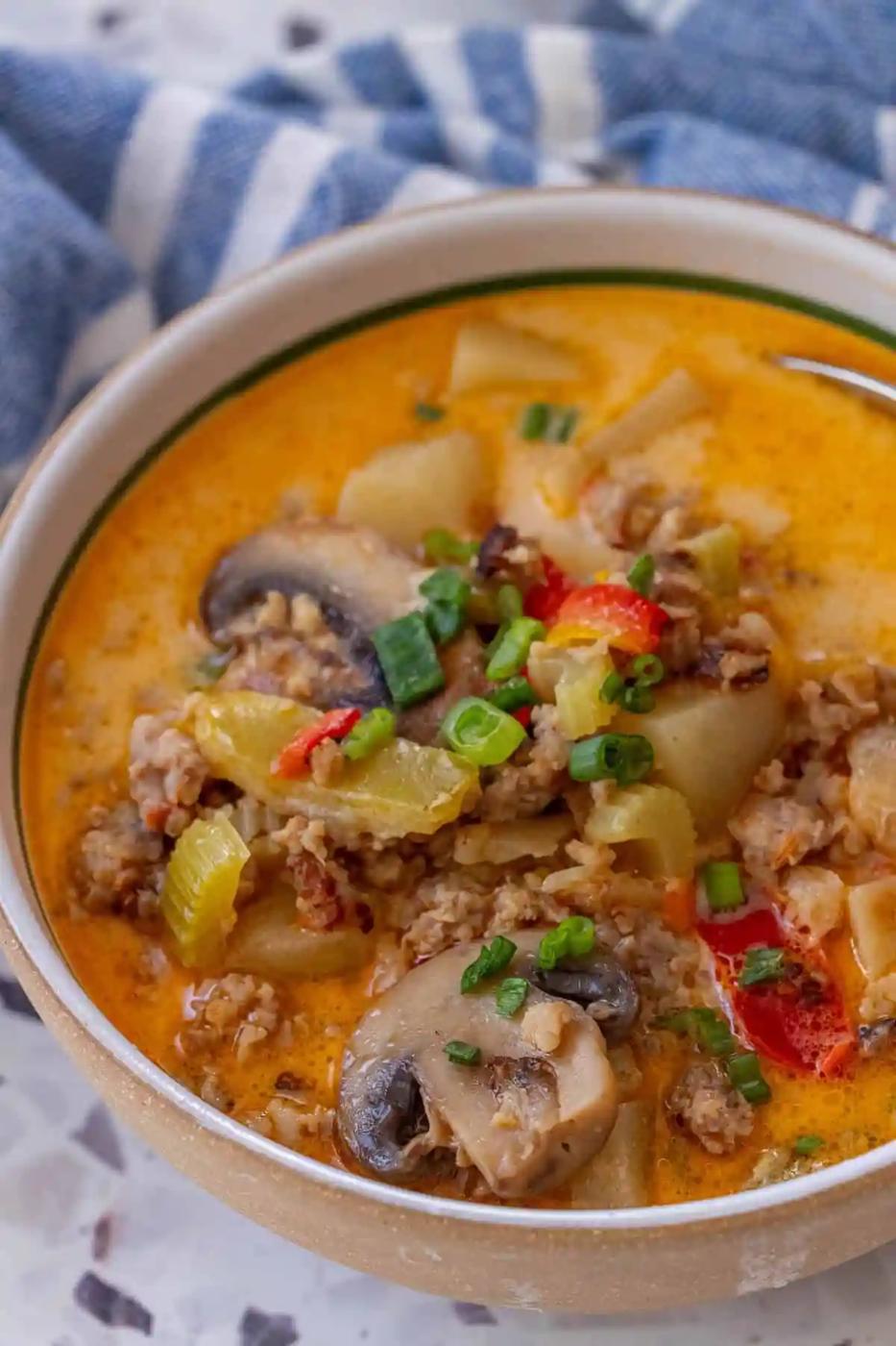 A bowl of 20 Minute Creamy Sausage and Mushroom Soup with sliced mushrooms, ground meat, potatoes, veggies, and green onions sits on a marble surface near a blue and white cloth.
