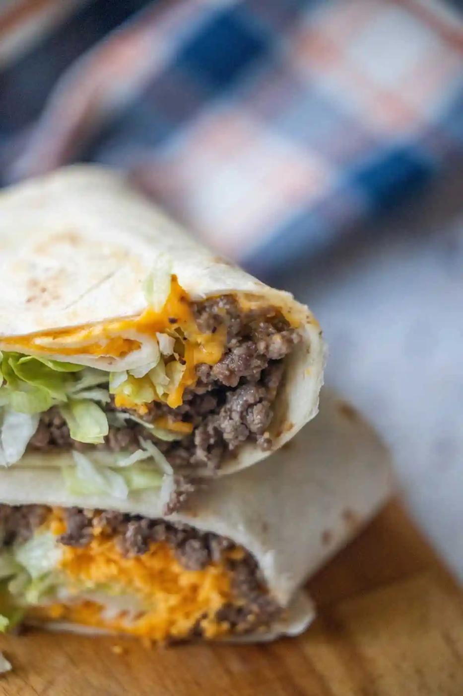A close-up of easy cheeseburger wraps cut in half, filled with ground beef, shredded cheddar cheese, and lettuce, stacked on a wooden surface.