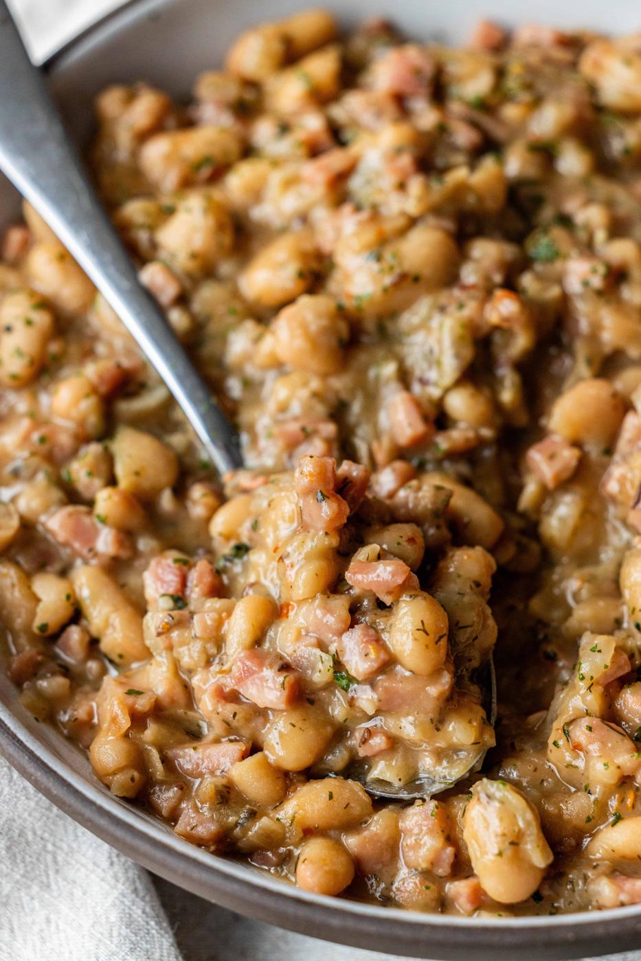 A close-up of a bowl of brothy ham and beans with diced ham, herbs, and a hint of caraway, with a spoon scooping up a portion.