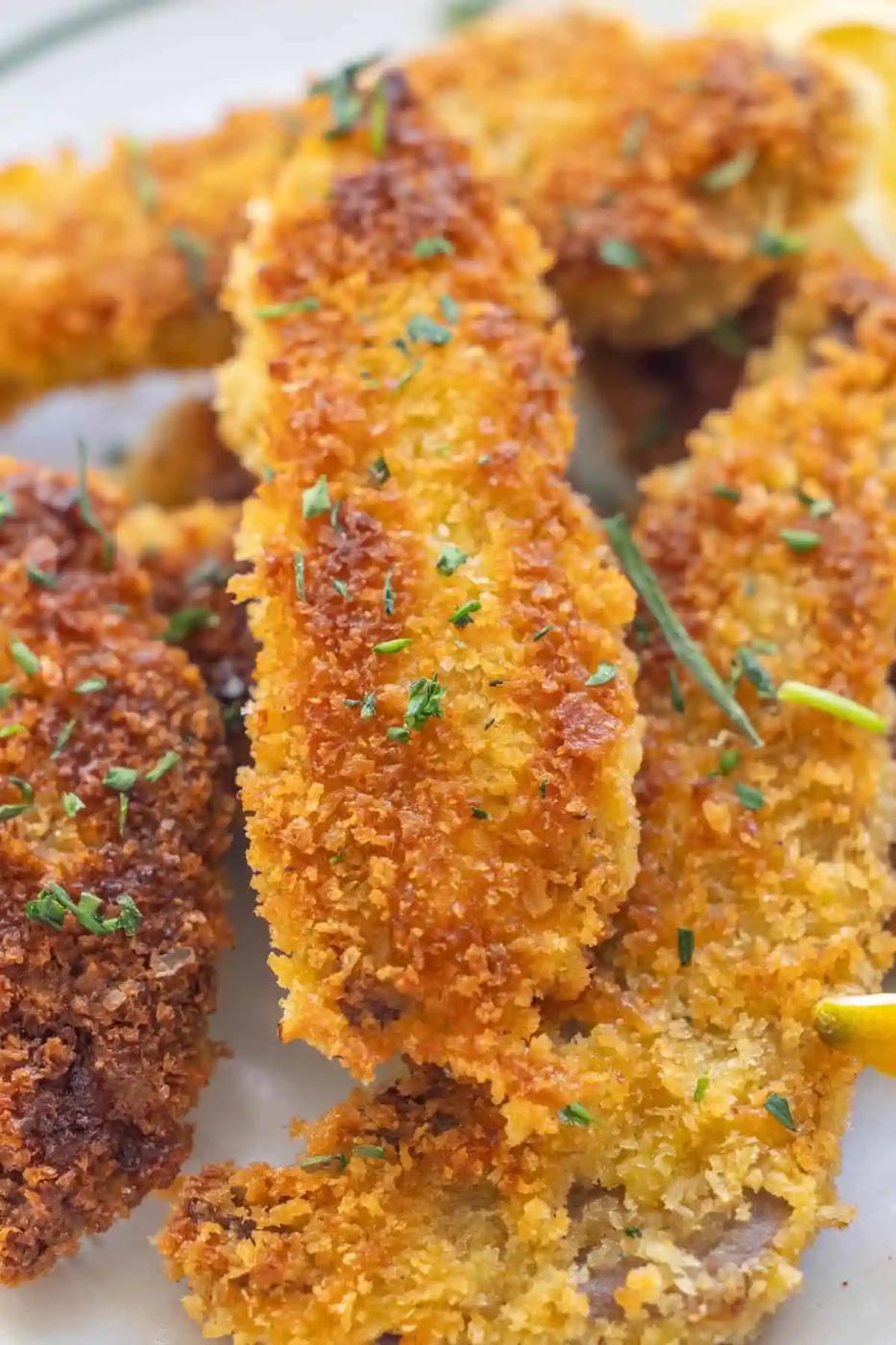 Close-up of several breaded and fried fish fillets garnished with chopped herbs and lemon slices on a plate.