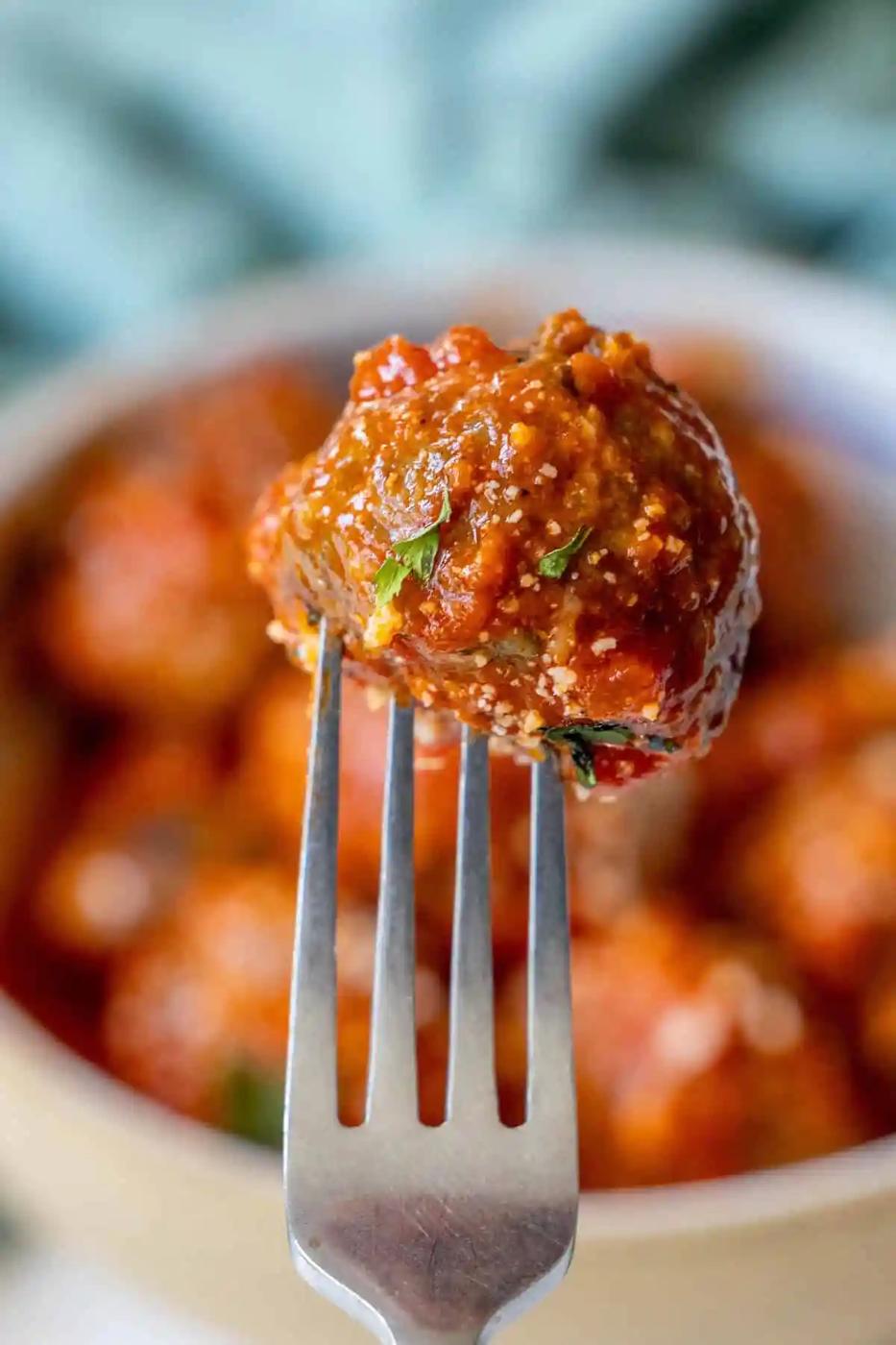 A close-up of a fork holding a meatball covered in tomato sauce with a blurred background of more baked meatballs.
