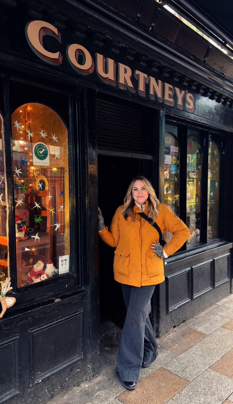 A person in a yellow jacket stands in the doorway of a black storefront labeled "Courtneys," reminiscent of Courtney O'Dell Ajpal, with decorated windows displaying various items.