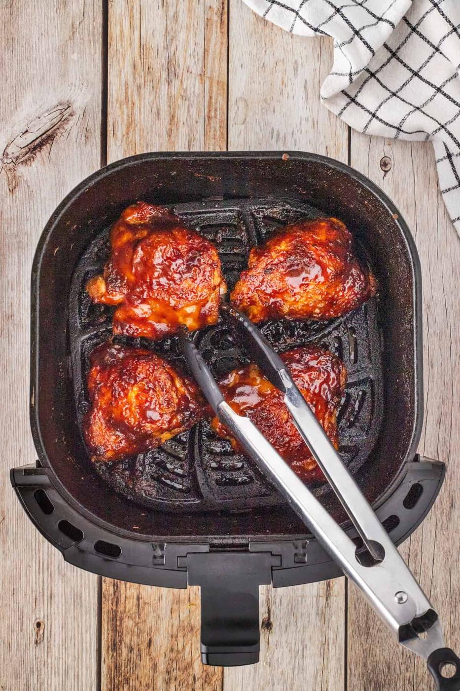 Three juicy pieces of glazed BBQ chicken thighs sit in an air fryer basket, with stainless steel tongs grasping one piece. A checkered towel is partially visible on the wooden surface beside the fryer.