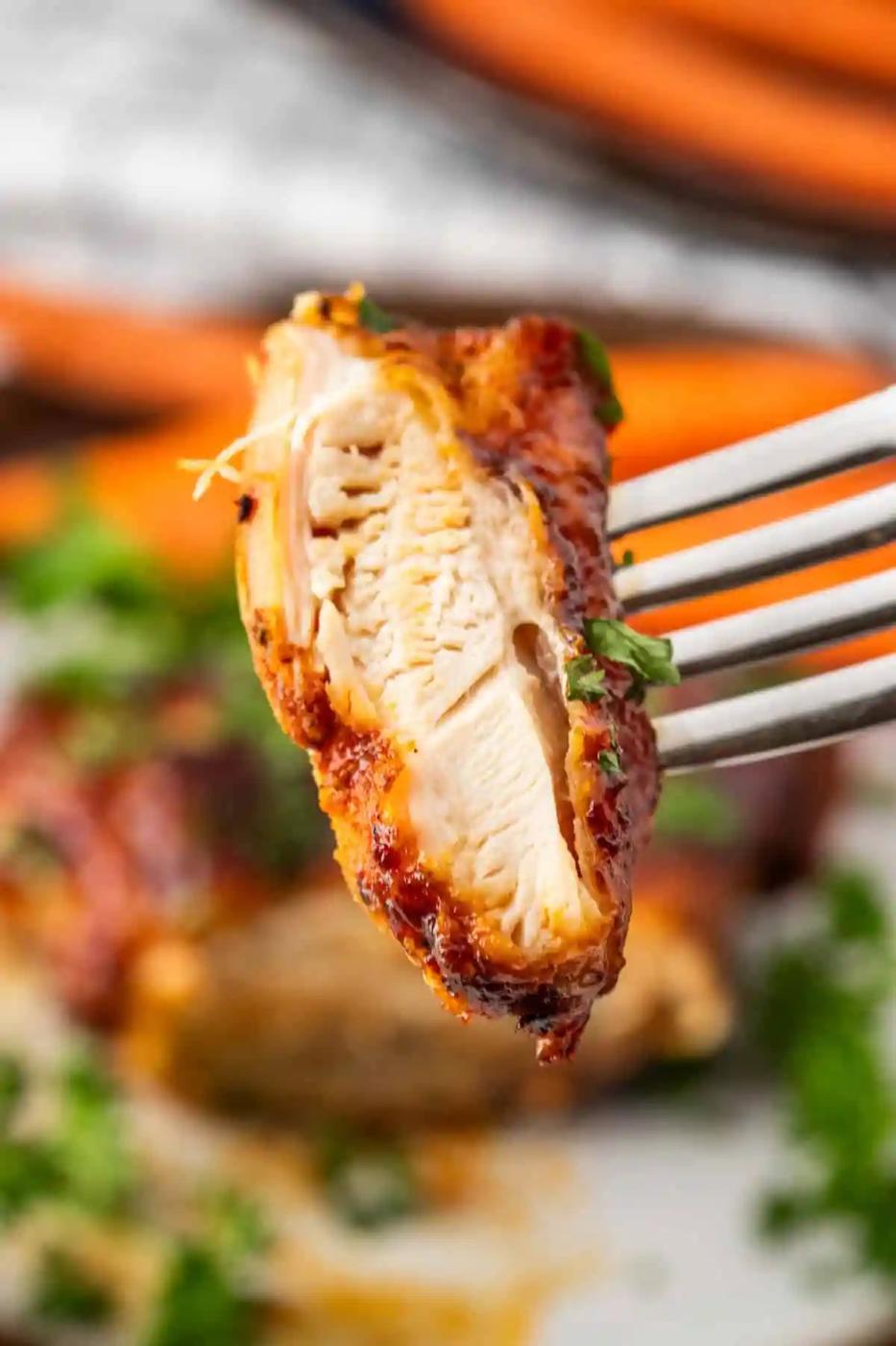 Close-up of a fork holding a piece of seasoned, cooked BBQ chicken with visible grill marks, against a blurred background of more succulent chicken thighs and green garnish.