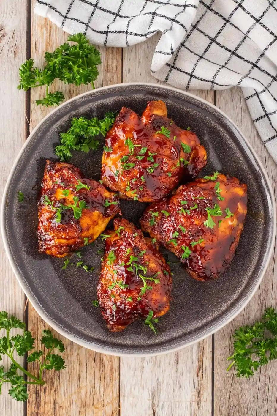 Four succulent pieces of glazed chicken thighs with chopped parsley rest on a dark plate, set on a wooden surface. Accompanied by a checkered cloth and fresh parsley sprigs, they're perfect for a quick meal straight from the air fryer.