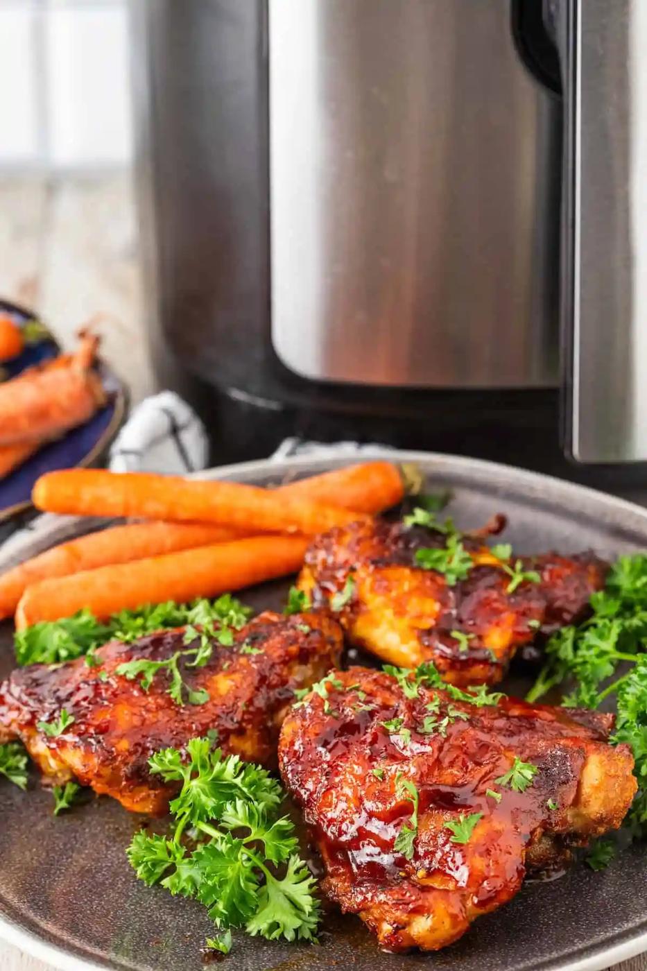 A plate of succulent BBQ chicken thighs garnished with parsley, accompanied by two whole carrots, sits invitingly. An air fryer hums softly in the background, ready for your next culinary creation.