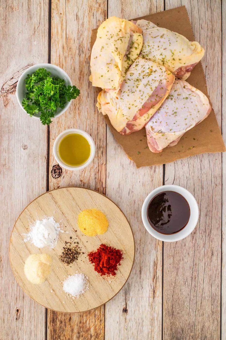 Raw chicken thighs, fresh parsley, and olive oil sit enticingly on a round wooden board surrounded by various spices on a rustic wooden table, ready to be transformed into succulent BBQ chicken in the air fryer.