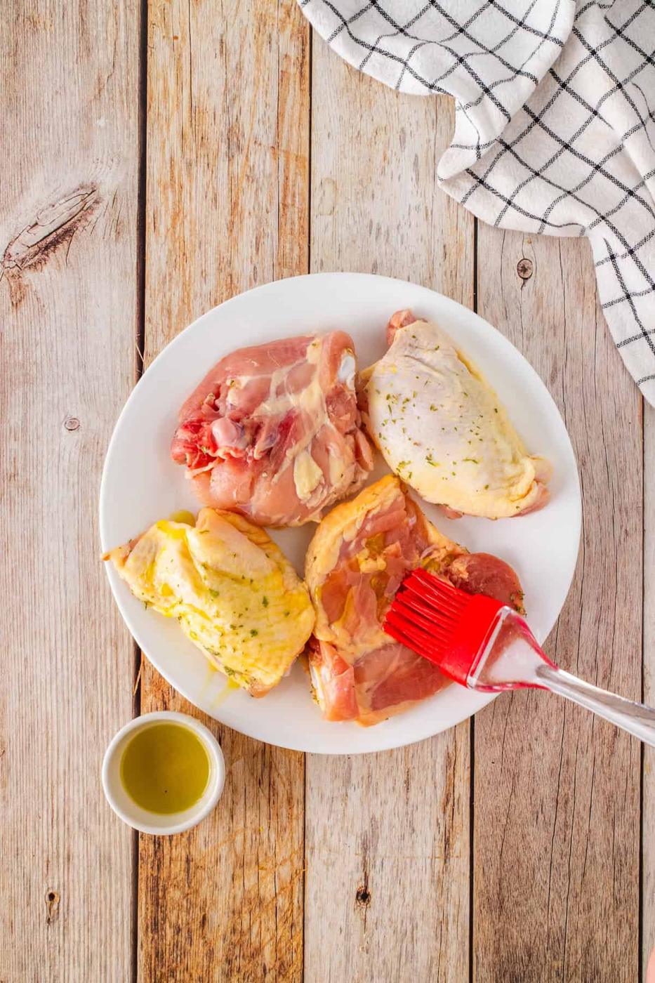 Raw chicken thighs on a white plate are being brushed with tangy BBQ marinade. A small bowl of marinade sits nearby. The plate rests on a wooden surface with a checkered cloth beside it, ready for the air fryer magic.