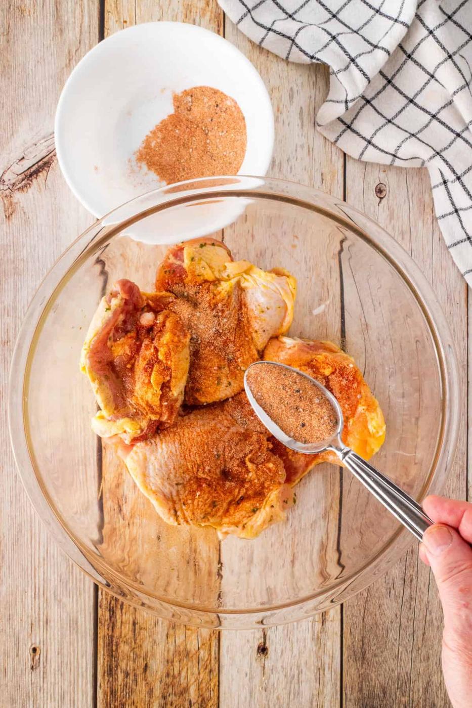 Chicken thighs in a glass bowl are being seasoned with a spice mix. A hand holds a spoon, blending flavors. Nearby, a small white bowl with extra seasoning rests on a wooden surface adorned with a checkered cloth. Perfect for air fryer BBQ chicken indulgence!.