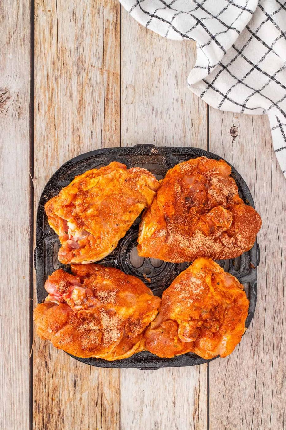 Four seasoned chicken thighs rest on a black grill pan, ready to become delicious BBQ chicken, placed on a wooden surface with a checkered cloth nearby.