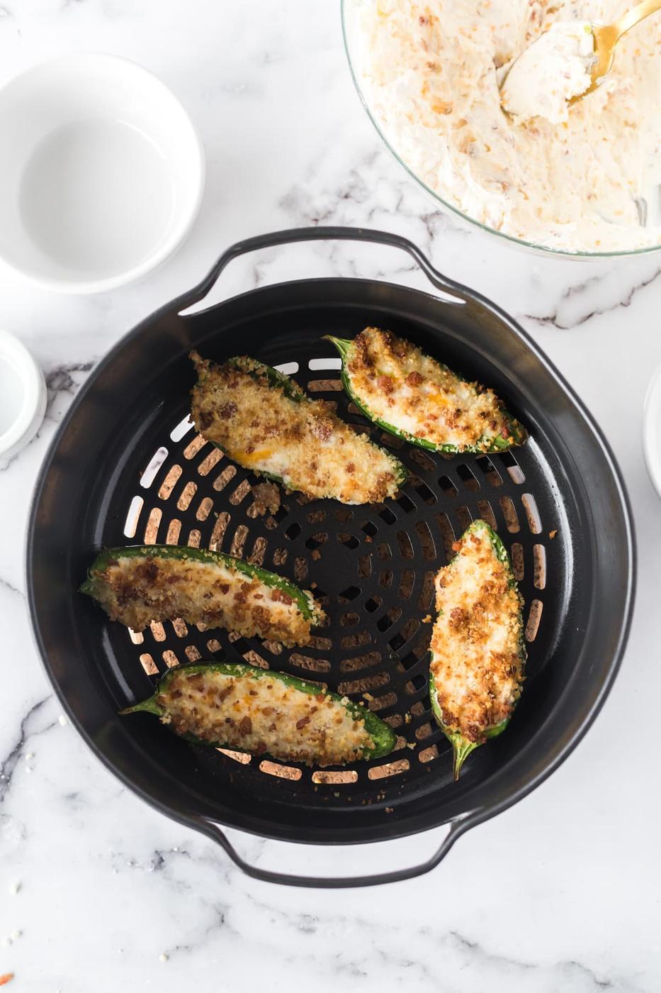 browned air fried jalapeño peppers cut in half filled with cream cheese filling and topped with panko breadcrumbs in the basket of an air fryer