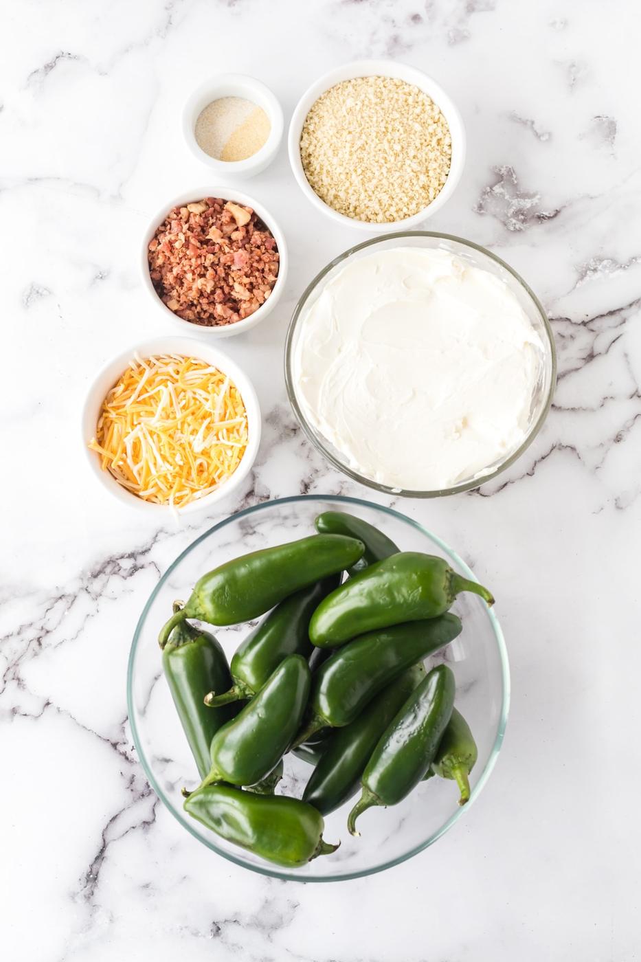 jalapeño peppers in a bowl, cream cheese in a bowl, cheese in a bowl, crumbled bacon in a bowl, breadcrumbs in a bowl, garlic salt and onion powder in a bowl on a table