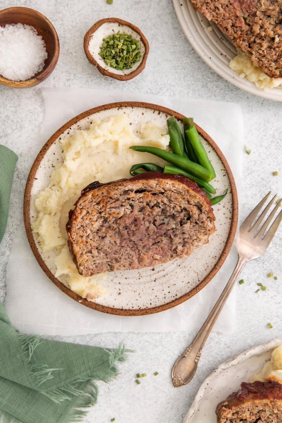 picture of a slice of meatloaf on a white plate on a table next to a fork