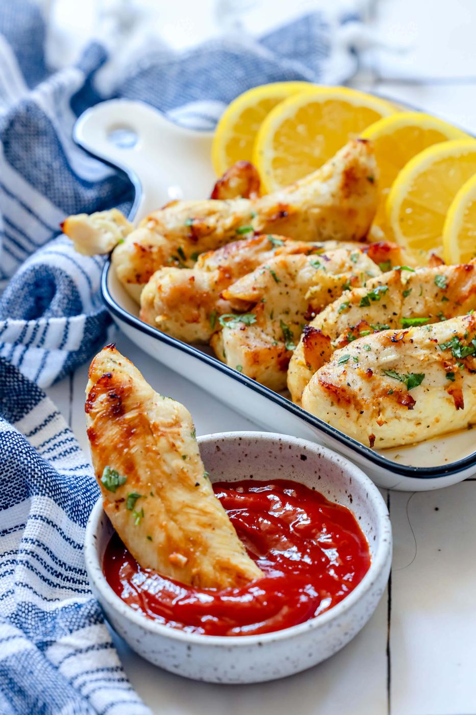 naked chicken tenders in a white dish with lemon slices next to them and chopped parsley on top