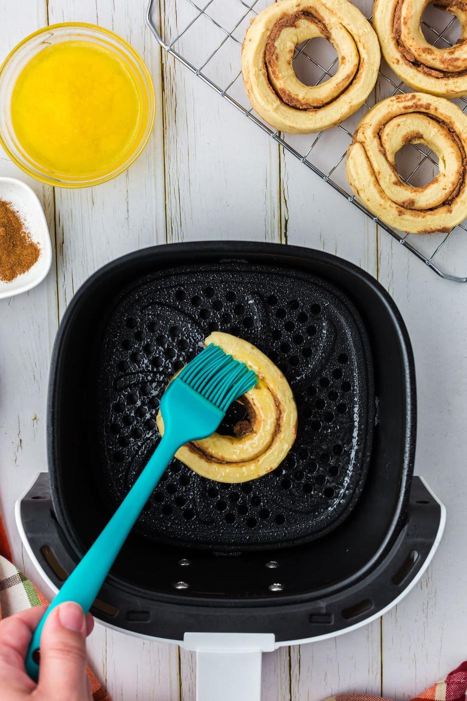 a silicon spatula brushing melted butter on a cinnamon roll donut in an air fryer basket