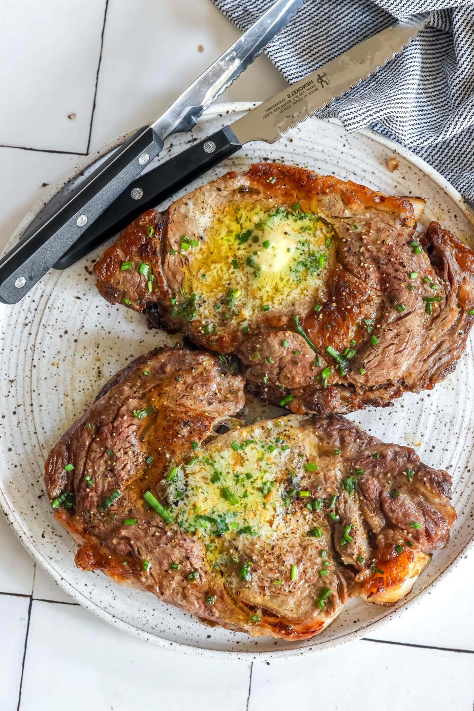Two pan fried ribeye steaks on a plate with herbs and a knife.