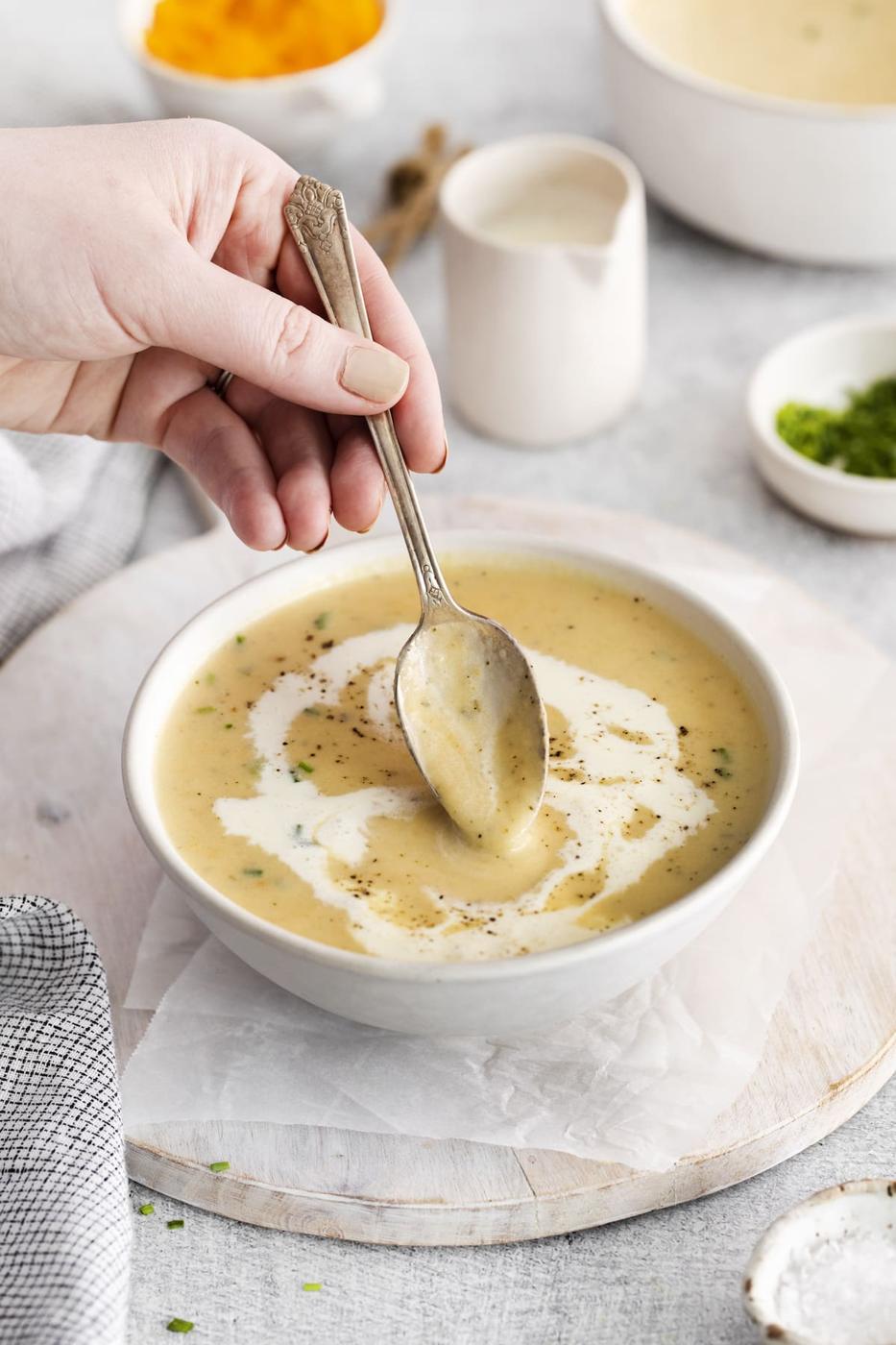 a hand holding a spoon in a bowl of cheddar soup with a swirl of cream and crack of pepper on top