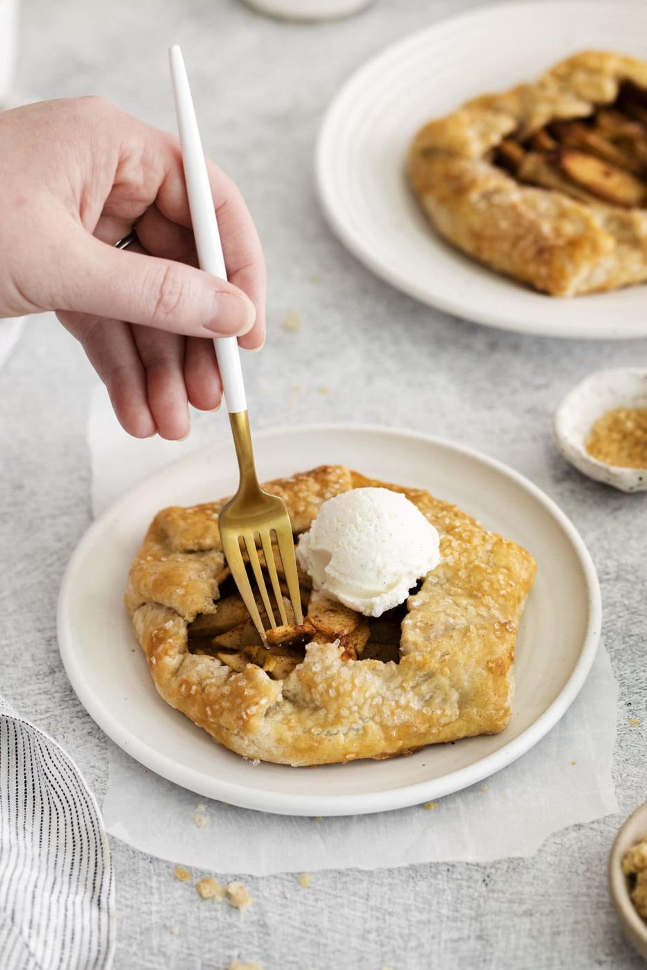 picture of apple galette on a table with a scoop of ice cream and hand holding a fork cutting into it