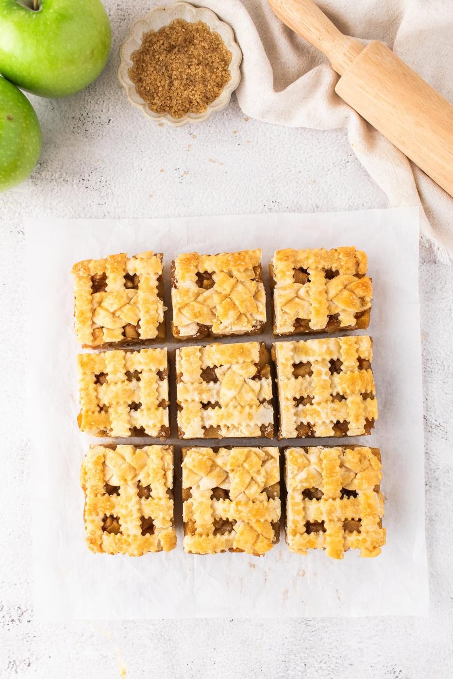 Nine square Apple Pie Bars with lattice tops sit on parchment paper, surrounded by two green apples, brown sugar, and a rolling pin for a cozy homemade touch.