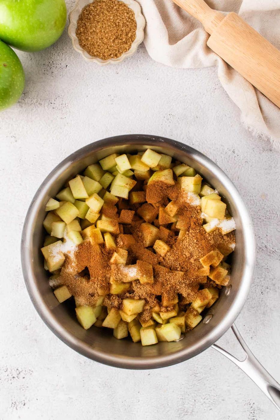 A saucepan filled with diced green apples, brown sugar, granulated sugar, and cinnamon—perfect for Apple Pie Bars—sits beside a rolling pin, sugar, and whole apples on a light countertop.