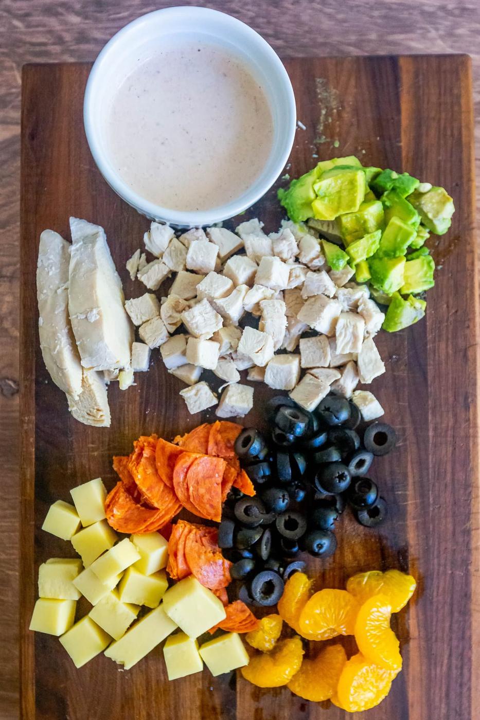 picture of cooked chicken breast, avocado, pepperoni, cheese, olives, and mandarin oranges on a cutting board