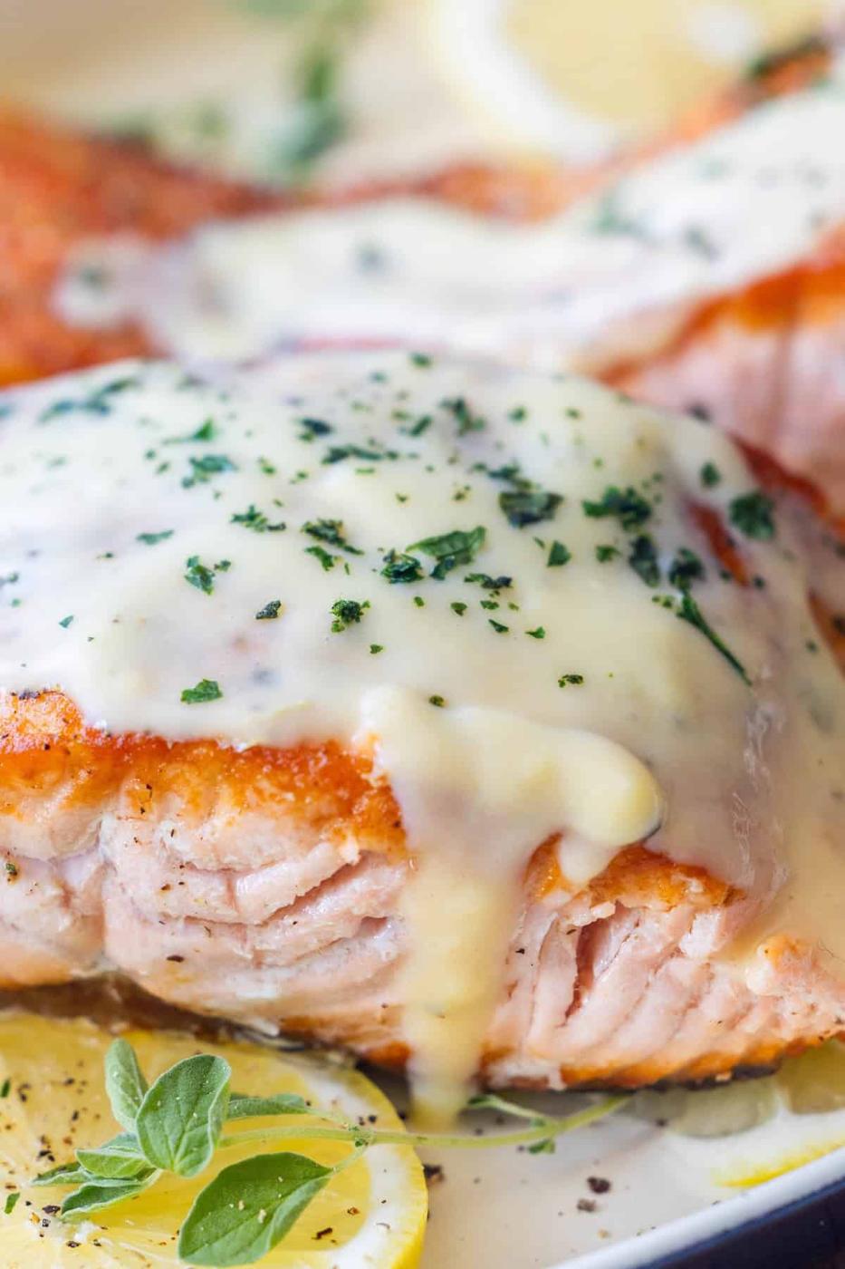 Close-up of cooked Greek salmon fillet topped with creamy white sauce and chopped herbs, garnished with lemon slices and oregano—an irresistible Greek salmon recipe.