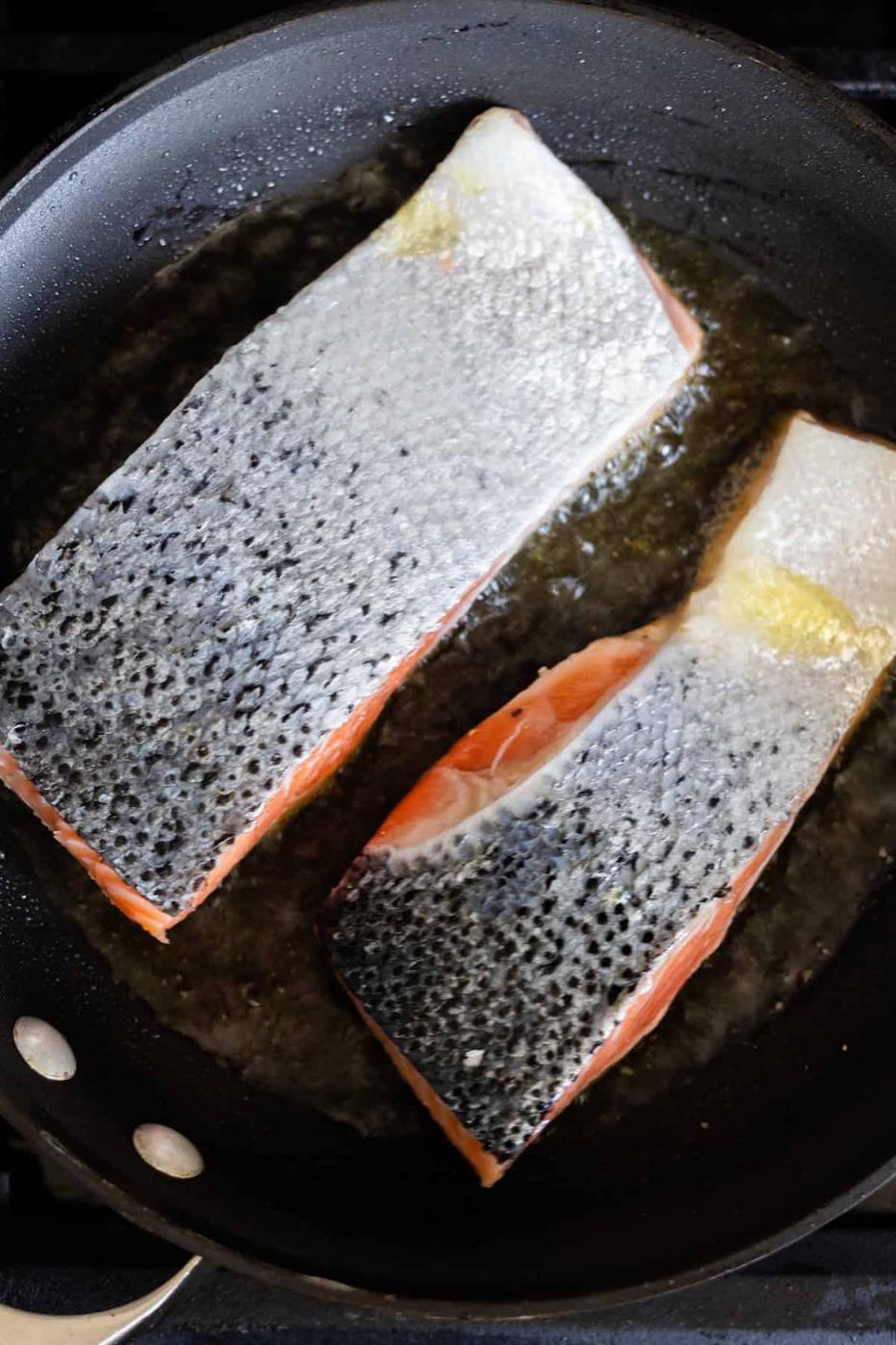 Two raw Pan Fried Salmon Filets, skin side down, sizzle in a black frying pan with oil.