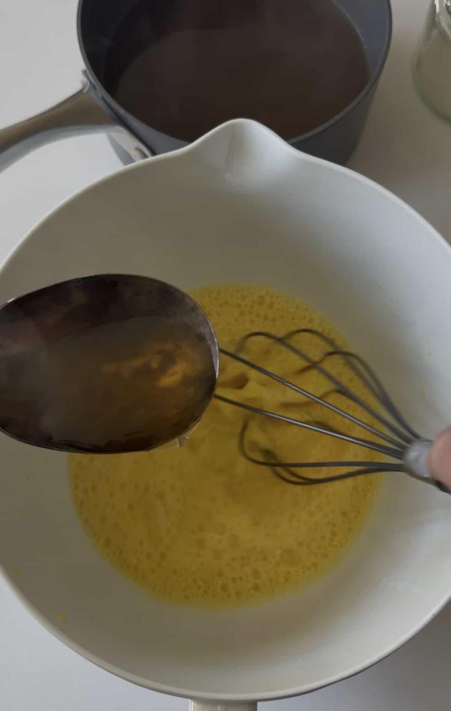 A metal spoon pours liquid into a bowl of beaten eggs being whisked, as Avgolemono sauce—a classic Greek sauce—comes together, with a saucepan and jar in the background.