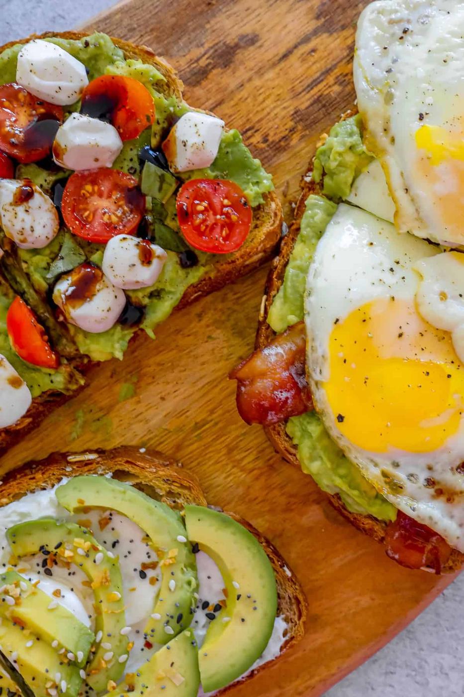 Four slices of toast with avocado, bacon, tomatoes and eggs on a wooden cutting board.