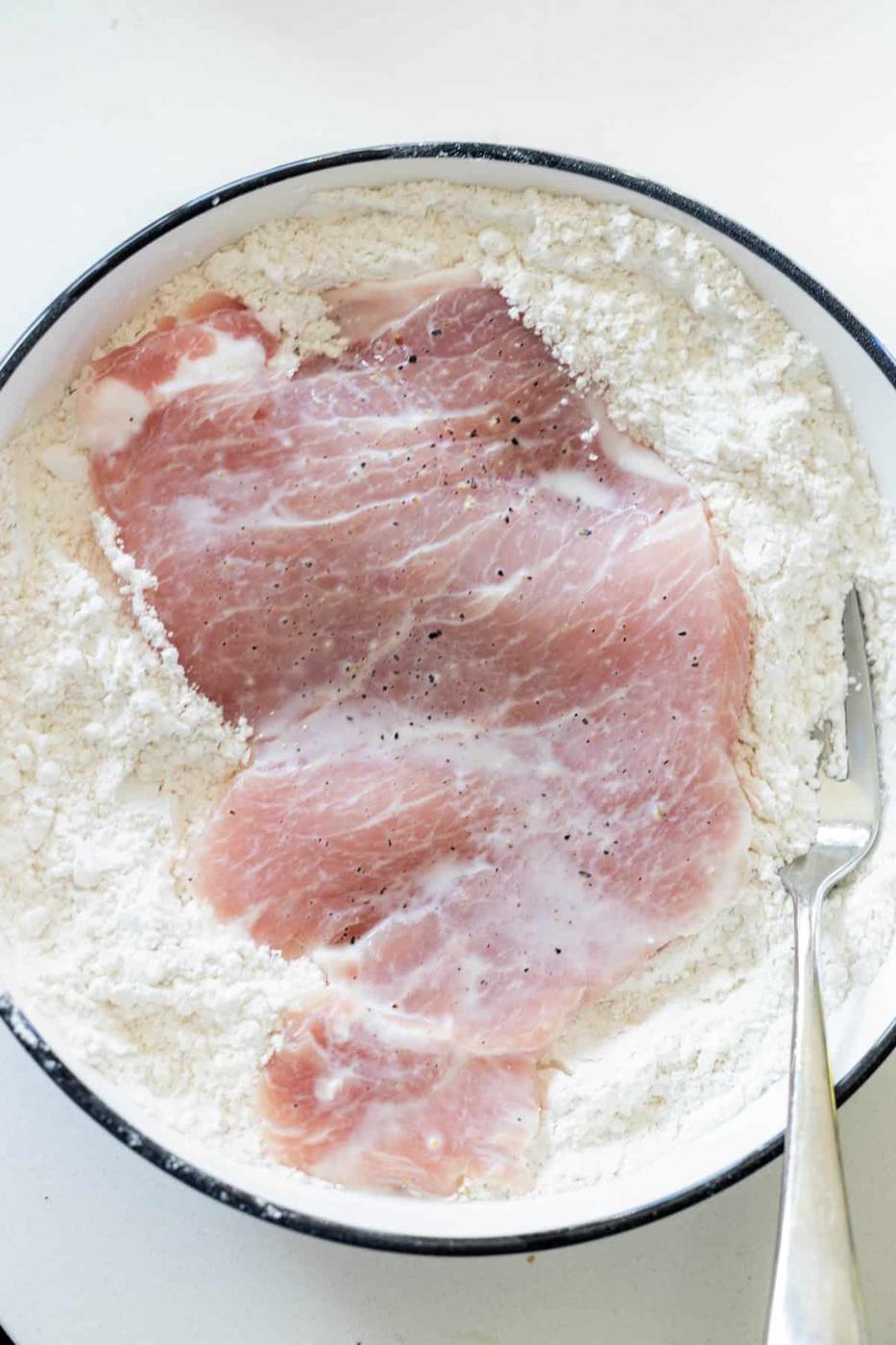 Raw pork cutlet coated with flour and pepper in a white shallow dish, ready to be transformed into crispy Breaded Pork Chops, with a metal fork resting at the edge.
