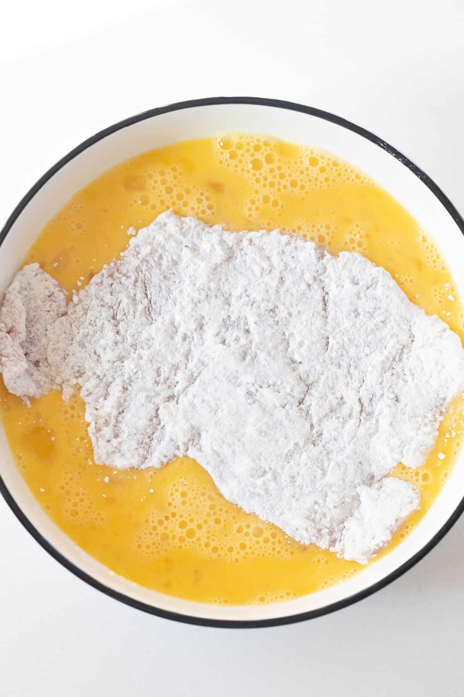 A piece of flour-coated meat is dipped in a bowl of beaten eggs, a classic step when making breaded pork chops before baking them to perfection.