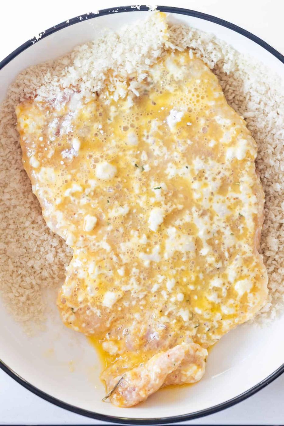 Raw chicken breast coated in beaten egg is placed on a layer of panko breadcrumbs in a white bowl, ready for breading—just like preparing Breaded Pork Chops or Baked Pork Chops.