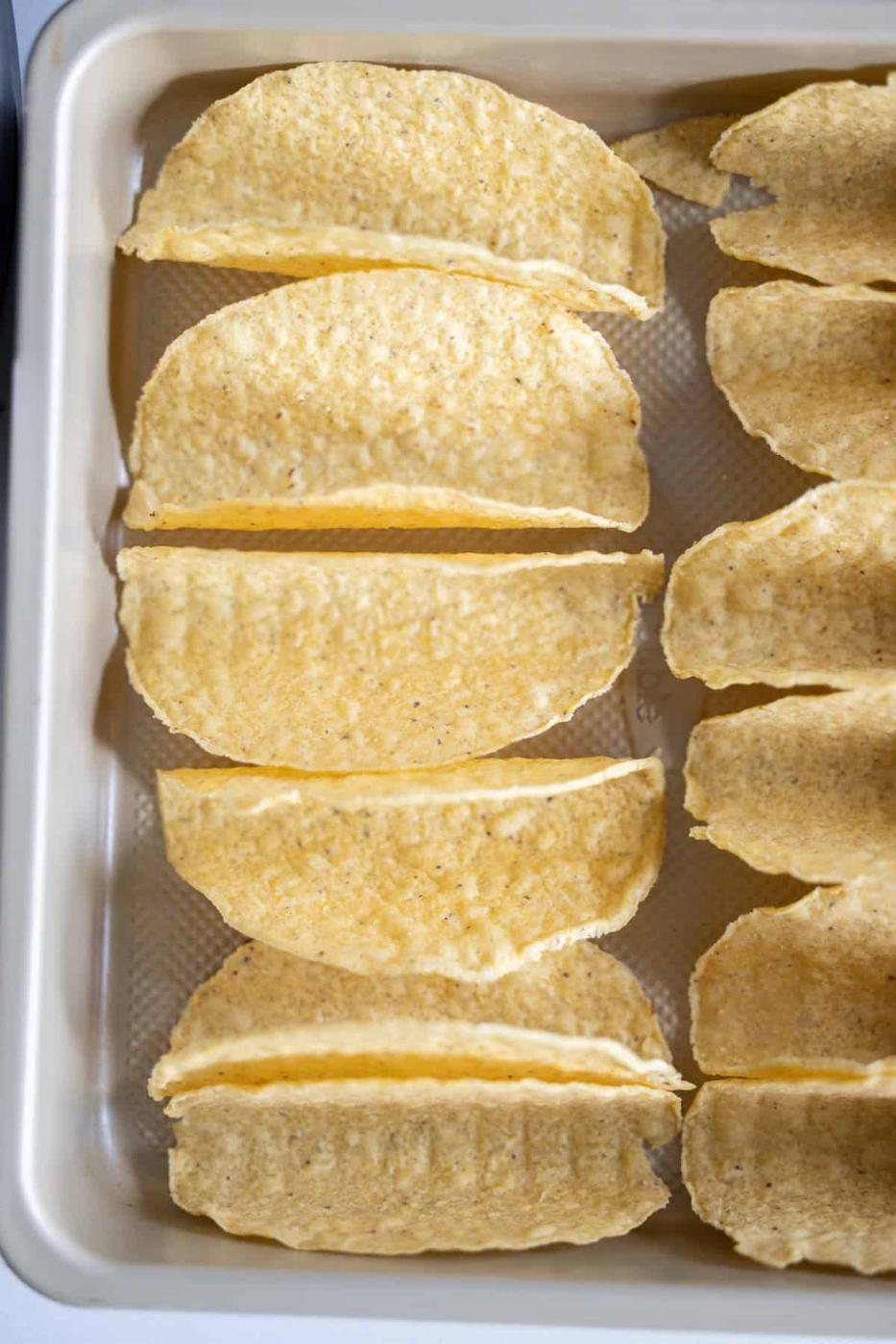 A baking tray lined with upright, empty hard taco shells arranged in rows, ready to be filled for delicious Baked Chicken Tacos.