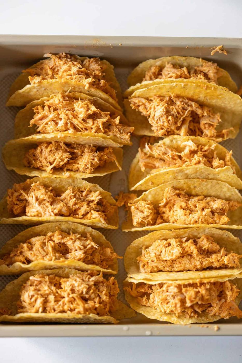 A baking tray holds rows of Baked Chicken Tacos, each hard shell filled with shredded, seasoned chicken, arranged neatly for baking or serving.