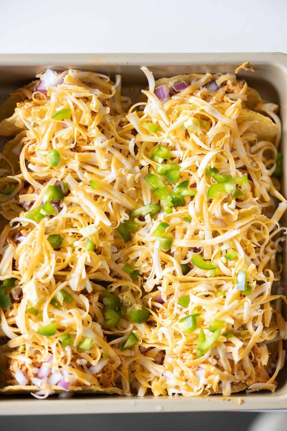 A baking dish filled with tortilla chips, shredded cheese, chopped green bell peppers, and red onions—just like the perfect Baked Chicken Tacos, ready to be baked to melty perfection.