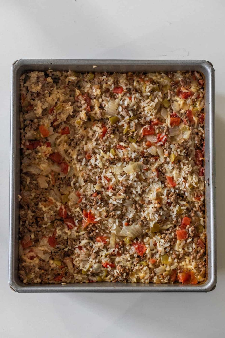 A square baking pan filled with Baked Dirty Rice casserole, featuring ground meat, rice, chopped onions, red bell peppers, and green bell peppers.