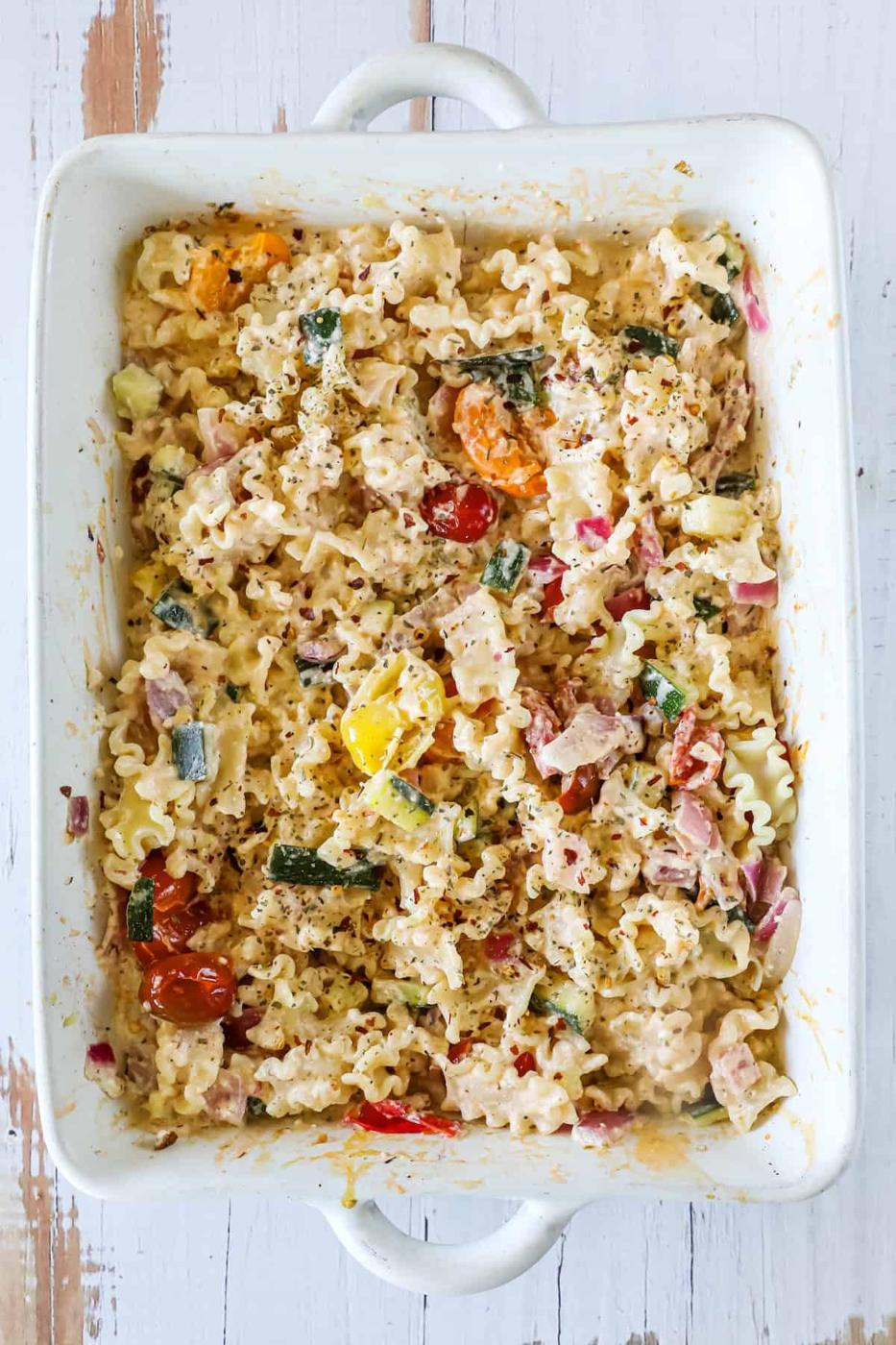 A rectangular casserole dish filled with cooked pasta, tomatoes, zucchini, and herbs mixed with a creamy sauce.