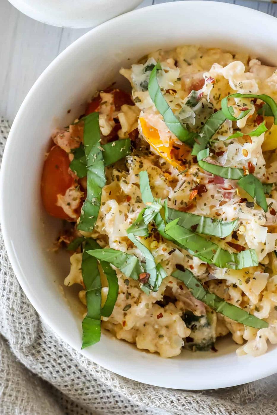 A white bowl filled with creamy macaroni and cheese, topped with chopped fresh basil and a mix of red and yellow cherry tomatoes.